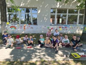 Grupa dzieci z oddziału przedszkolnego pozuje do wspólnego zdjęcia na tle przezroczystej folii ozdobionej kolorowymi pracami plastycznymi. Fotografia została wykonana podczas pikniku z okazji Dnia Dziecka na terenie szkoły.