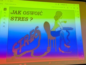 Slajd prezentacji wyświetlony na ekranie z tytułem „Jak oswoić stres?”. Grafika przedstawia siedzącą przy stoliku postać oraz stylizowany napis „stres” w formie przewodu elektrycznego.