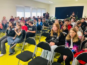 Uczniowie klas starszych siedzą na krzesłach w szkolnej sali konferencyjnej przed rozpoczęciem spotkania poświęconego radzeniu sobie ze stresem. W tle widoczni są nauczyciele i organizatorzy wydarzenia.
