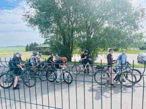 Grupa uczniów z rowerami i kaskami gromadzi się przed rozpoczęciem rajdu rowerowego. Uczestnicy stoją przy swoich rowerach na terenie szkoły, w tle widoczne są drzewa oraz otwarta przestrzeń wiejska.