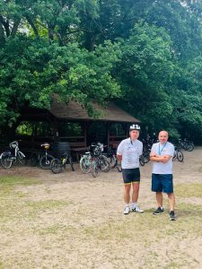 Dwóch dorosłych uczestników akcji rowerowej stoi przed drewnianą wiatą na Barbarce. Wokół znajdują się zaparkowane rowery, a miejsce otacza zielony las.