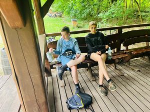 Dwoje uczniów odpoczywa na ławce pod drewnianą wiatą na Barbarce. Młodzież korzysta z przerwy podczas rajdu rowerowego, a w tle widoczne są rowery i leśne otoczenie.