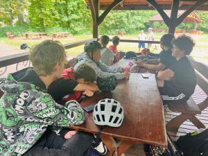 Grupa uczniów siedzi przy drewnianym stole pod wiatą podczas postoju na Barbarce. Na stole leżą kaski i napoje, a uczestnicy odpoczywają po przejechanym odcinku trasy.