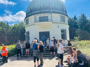 Grupa uczniów i opiekunów zgromadzona przed budynkiem obserwatorium astronomicznego z charakterystyczną kopułą. Uczestnicy oczekują na rozpoczęcie zwiedzania.