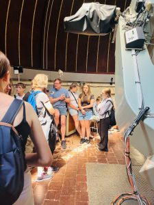 Wnętrze kopuły obserwatorium. Przewodnik opowiada uczniom o działaniu teleskopu, a uczestnicy słuchają wyjaśnień i obserwują wyposażenie naukowe.