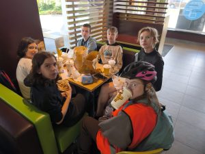 Grupa uczniów siedzi przy stoliku w restauracji podczas przerwy na posiłek. Na stole znajdują się napoje i zamówione zestawy obiadowe.