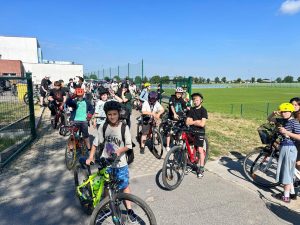 Uczniowie na rowerach oczekują na rozpoczęcie wycieczki rowerowej. Kilkanaście osób ustawiło się przy wyjeździe z terenu szkoły. Większość uczestników ma kaski ochronne, a w tle widoczne jest boisko sportowe oraz otwarta przestrzeń wokół szkoły.