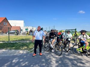 Nauczyciel wychowania fizycznego wraz z grupą uczniów na rowerach przygotowują się do wyjazdu spod szkoły. Uczestnicy stoją na drodze przy szkolnym ogrodzeniu, a część uczniów siedzi już na rowerach. Zdjęcie wykonano w słoneczny dzień podczas akcji promującej aktywność rowerową.
