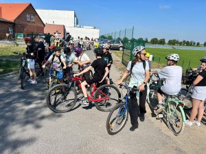 Grupa uczniów oraz opiekunowie na rowerach gromadzą się przed rozpoczęciem przejazdu. Uczestnicy rozmawiają i przygotowują sprzęt do jazdy. W tle widoczne są budynki szkoły, zaparkowane samochody oraz boisko sportowe.
