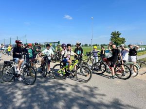 Uczniowie i opiekunowie na rowerach pozują do zdjęcia przed wyjazdem na trasę rowerową. Kilku uczestników macha do fotografa. Wszyscy znajdują się na terenie przy szkole, obok boiska sportowego. Zdjęcie dokumentuje udział społeczności szkolnej w akcji „Rowerowa Stolica Polski”.