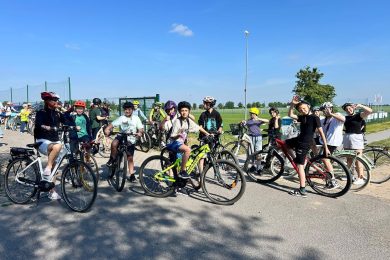 Uczniowie i opiekunowie na rowerach pozują do zdjęcia przed wyjazdem na trasę rowerową. Kilku uczestników macha do fotografa. Wszyscy znajdują się na terenie przy szkole, obok boiska sportowego. Zdjęcie dokumentuje udział społeczności szkolnej w akcji „Rowerowa Stolica Polski”.