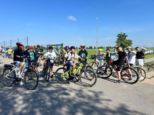 Uczniowie i opiekunowie na rowerach pozują do zdjęcia przed wyjazdem na trasę rowerową. Kilku uczestników macha do fotografa. Wszyscy znajdują się na terenie przy szkole, obok boiska sportowego. Zdjęcie dokumentuje udział społeczności szkolnej w akcji „Rowerowa Stolica Polski”.