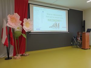 Uczennica siedzi przy stanowisku komputerowym i obsługuje prezentację wyświetlaną podczas spotkania edukacyjnego poświęconego radzeniu sobie ze stresem. W tle widoczny jest ekran projekcyjny oraz dekoracje sali.
