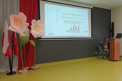 Uczennica siedzi przy stanowisku komputerowym i obsługuje prezentację wyświetlaną podczas spotkania edukacyjnego poświęconego radzeniu sobie ze stresem. W tle widoczny jest ekran projekcyjny oraz dekoracje sali.