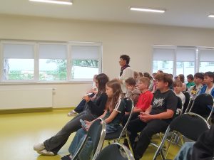 Uczniowie uczestniczą w spotkaniu edukacyjnym dotyczącym stresu. Prelegentka rozmawia z młodzieżą, która aktywnie słucha i obserwuje prezentowane treści.