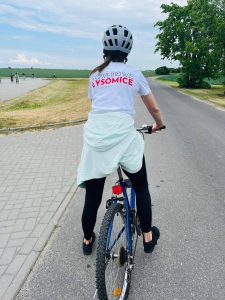 Uczestniczka rajdu rowerowego jedzie drogą wśród pól. Na koszulce widoczny jest napis „Rowerowe Łysomice”. W oddali można zauważyć innych rowerzystów poruszających się trasą wycieczki.