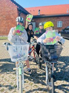 Czworo uczniów na rowerach prezentuje książki zabrane na rajd rowerowy. Uczestnicy w kaskach stoją przed ceglanym budynkiem szkolnym, trzymając książki w dłoniach i uśmiechając się do zdjęcia.