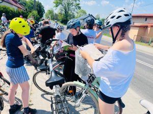 Uczniowie podczas przerwy w rajdzie rowerowym stoją przy swoich rowerach i czytają książki. Zdjęcie wykonano przy drodze w słoneczny dzień. Widoczne są rowery, kaski ochronne oraz uczestnicy przygotowujący się do dalszej jazdy.
