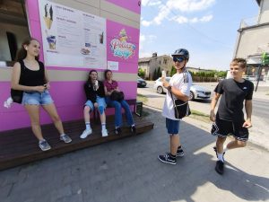 Grupa uczniów odpoczywa przy lodziarni podczas wycieczki rowerowej. Część uczestników siedzi na ławce, inni stoją z lodami w dłoniach. W tle znajduje się kolorowa ściana z menu lodziarni.