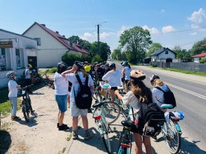 Grupa uczestników wycieczki rowerowej zatrzymała się przy drodze na krótki odpoczynek. Uczniowie stoją obok rowerów, rozmawiają i przygotowują się do dalszej jazdy. W tle widoczne są domy jednorodzinne oraz drzewa rosnące wzdłuż ulicy.