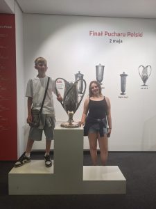 Dwójka uczniów pozuje przy trofeum Pucharu Polski prezentowanym na ekspozycji Stadionu Narodowego. W tle znajdują się informacje o historii rozgrywek.