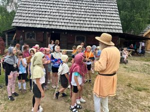 Przewodnik w stroju z epoki opowiada dzieciom o życiu w średniowieczu. Uczniowie stoją na dziedzińcu grodu w kolorowych nakryciach głowy nawiązujących do dawnych strojów.