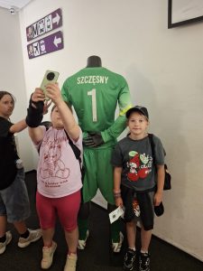 Uczennica wykonuje zdjęcie przy ekspozycji z koszulką bramkarską Wojciecha Szczęsnego. Obok stoi koleżanka, a pozostali uczestnicy zwiedzają wystawę.