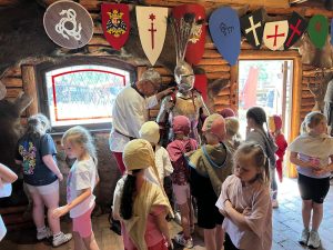 Zwiedzanie zbrojowni. Dzieci oglądają średniowieczną zbroję rycerską prezentowaną przez przewodnika. Na ścianach wiszą tarcze heraldyczne i elementy wyposażenia wojowników.