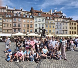 Grupa uczniów wraz z opiekunami pozuje do wspólnego zdjęcia na Rynku Starego Miasta w Warszawie. W tle znajduje się Pomnik Syrenki Warszawskiej oraz kolorowe kamienice.