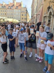 Uczniowie prezentują zakupione pamiątki podczas spaceru po warszawskiej Starówce. W tle widoczni są pozostali turyści i zabytkowe kamienice.