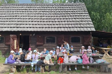 Przerwa podczas wycieczki. Uczniowie siedzą przy drewnianych ławach przed budynkiem grodu, spożywają posiłek i odpoczywają po warsztatach oraz zwiedzaniu.