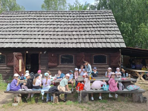 Przerwa podczas wycieczki. Uczniowie siedzą przy drewnianych ławach przed budynkiem grodu, spożywają posiłek i odpoczywają po warsztatach oraz zwiedzaniu.