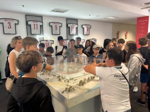 Uczniowie słuchają przewodnika podczas zwiedzania Stadionu Narodowego. W centrum znajduje się makieta stadionu, a na ścianie wiszą koszulki reprezentacji Polski.