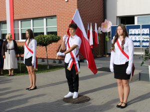 Poczet flagowy z biało-czerwoną flagą podczas uroczystości rozpoczęcia roku szkolnego przed budynkiem szkoły.