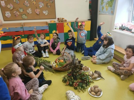 Uczniowie siedzą w kręgu wokół jesiennej ekspozycji z warzywami, ziemniakami, orzechami i roślinami podczas Święta Pieczonego Ziemniaka.