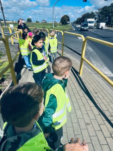 Dzieci wraz z nauczycielem obserwują ruch drogowy i uczą się zasad bezpiecznego zachowania w pobliżu jezdni.