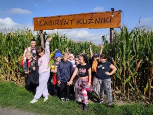 Uczniowie klas II i III wraz z opiekunką pozują przed wejściem do kukurydzianego labiryntu pod tablicą z napisem „Labirynt Kuźniki”.