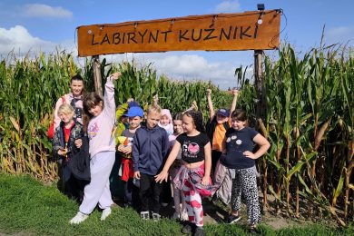 Uczniowie klas II i III wraz z opiekunką pozują przed wejściem do kukurydzianego labiryntu pod tablicą z napisem „Labirynt Kuźniki”.
