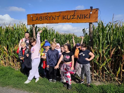 Uczniowie klas II i III wraz z opiekunką pozują przed wejściem do kukurydzianego labiryntu pod tablicą z napisem „Labirynt Kuźniki”.
