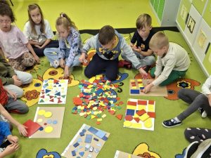 Uczniowie klasy I wspólnie pracują na dywanie z kolorowymi figurami geometrycznymi, układając je według różnych cech.