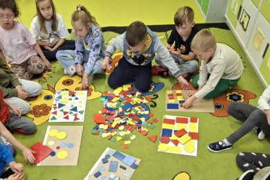 Uczniowie klasy I wspólnie pracują na dywanie z kolorowymi figurami geometrycznymi, układając je według różnych cech.