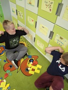 Dwóch uczniów klasy I siedzi na dywanie i układa kompozycje z kolorowych figur geometrycznych.