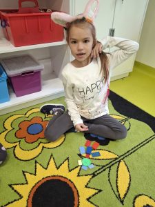 Uczennica klasy I siedzi na dywanie podczas zajęć matematycznych, przed nią znajdują się kolorowe figury geometryczne.