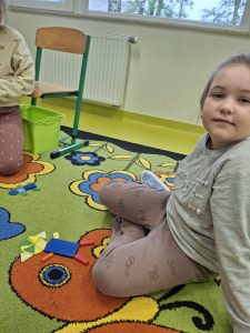 Uczennica klasy I podczas zajęć matematycznych siedzi na dywanie obok ułożonej z figur geometrycznych kompozycji.