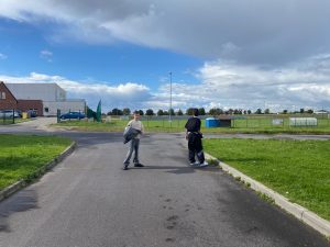 Dwóch uczniów z workami na odpady stoi na drodze przy boisku i budynku szkoły.