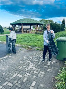 Dwóch uczniów trzyma worki na odpady na terenie szkoły, obok altany i zielonego pojemnika.