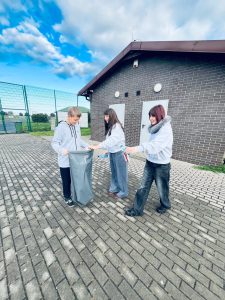 Troje uczniów zbiera odpady na utwardzonym terenie obok szkolnego budynku. Jeden z nich trzyma otwarty worek.