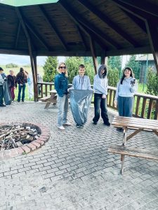 Nauczycielka i uczniowie stoją z workiem na odpady pod szkolną altaną. W tle inni uczniowie uczestniczą w sprzątaniu.