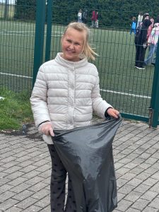 Uśmiechnięta uczennica w rękawiczce trzyma otwarty worek na odpady przy szkolnym boisku.