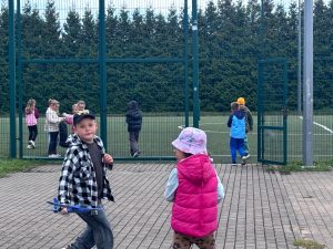 Uczniowie na terenie szkoły podczas akcji Sprzątania Świata. Część grupy znajduje się na boisku, a dwoje dzieci stoi przy otwartej furtce.